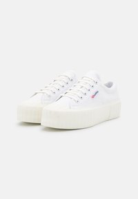Baskets en toile blanches avec une semelle épaisse et nervurée. Dotées d'un bout rond, d'un laçage à l'avant et d'un petit logo rouge et bleu sur le côté.