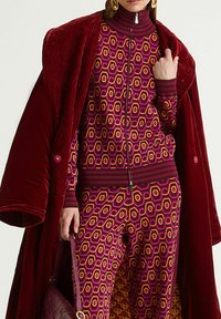 Ensemble de survêtement violet et rose à motifs hexagonaux avec un col zippé, associé à un manteau en peluche rouge profond et un sac à main.