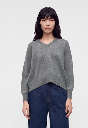 Frau mit langen schwarzen Haaren, die einen grauen V-Ausschnitt-Pullover und dunkelblaue Jeans trägt, vor einem einfarbigen hellen Hintergrund.