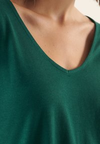 T-shirt verde a V in morbido tessuto; primo piano che mostra il collo e la texture liscia, senza motivi o accessori visibili.