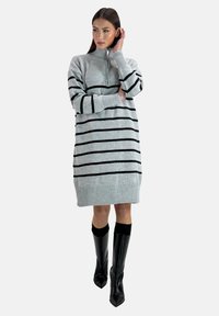 Robe en maille de couleur gris clair avec des rayures horizontales noires, col haut avec une fermeture éclaire, poignets et ourlet côtelés, portée avec des bottes noires allant jusqu'aux genoux.