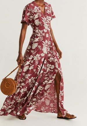 Femme portant une robe maxi fluide bordeaux à fleurs avec une fente latérale, portant un sac bandoulière rond marron et des sandales plates.