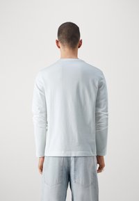 Mies, jolla on lyhyet hiukset ja joka pukeutuu yksinkertaiseen valkoiseen pitkähihaiseen paitaan ja vaaleansinisiin farkkuihin, seisoo selkä kameraan päin yksinkertaista taustaa vasten.