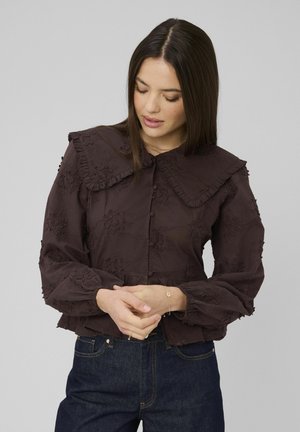 Femme portant un chemisier marron foncé brodé avec un grand col et des manches bouffantes, un jean bleu foncé, regardant vers le bas avec les mains jointes.