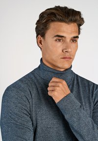 Niebieski sweter z golfem wykonany z dzianiny. Ma wysoki kołnierz oraz dopasowane mankiety, a także teksturowane wykończenie i subtelne, ciemne plamki.