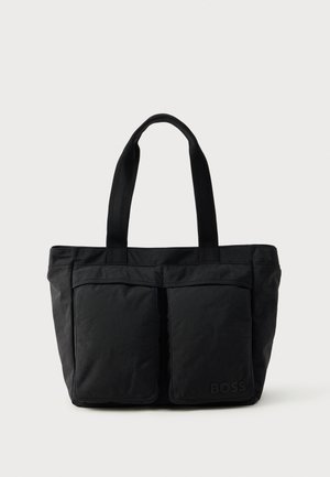 Black tote bag featuring a soft fabric construction, two front pockets, and sturdy handles. Logo displayed on the side. Simple, functional design.