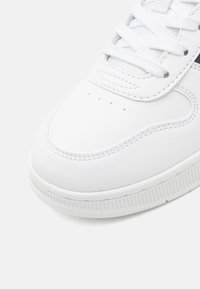 Hvit sneaker med glatt syntetisk materiale, rund tå, perforert design på tåboksen og teksturert gummiyttersåle. Har hvite lisser.