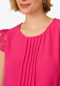 Top fucsia con maniche corte che presenta dettagli in pizzo intricati e pieghe verticali sul davanti; tessuto liscio e leggero.