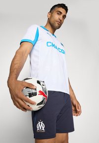Hvid fodboldtrøje med blå detaljer og logo, parret med marineblå shorts med et logo. Spiller holder en mønstret sort og hvid fodbold.