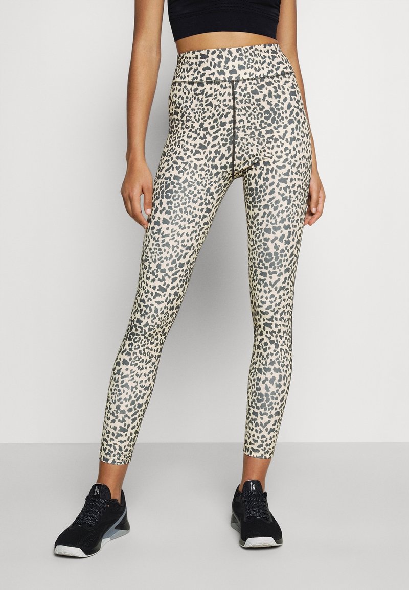 Leggings en imprimé léopard dans des tons beige et noir, taille haute avec une texture lisse, associés à des chaussures de sport noires.