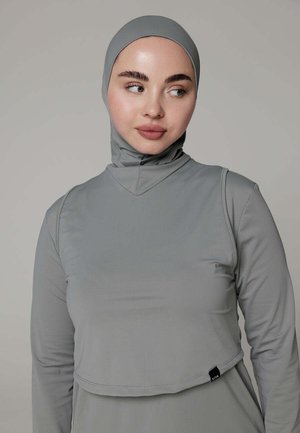 Vrouw die een aangesloten lichtgrijze hijab en bijpassende top met lange mouwen draagt, kijkt iets opzij tegen een effen achtergrond.