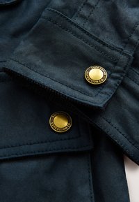 Marineblåt stof med en glat tekstur, der har to gyldne knapper med mærke og detaljerede syninger på ærmekanterne.