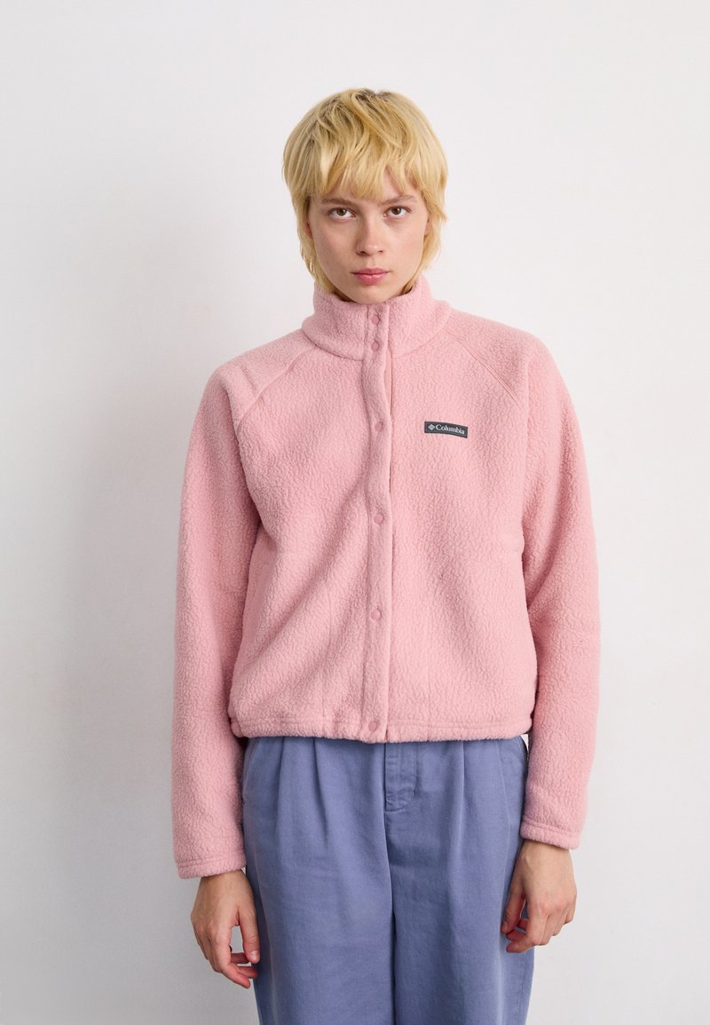 Persona con capelli biondi corti che indossa una giacca in pile rosa Columbia e pantaloni blu larghi su uno sfondo bianco semplice.