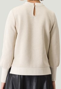 Beige langemouwen top met een schijnhalslijn, textuurstof en een keyhole-detail aan de achterkant. Bevat ribgebreide manchetten en zoom.