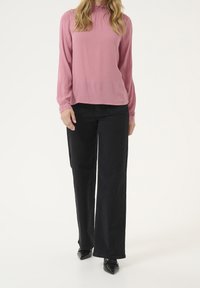 Blusa lunga a maniche di rosa chiaro con un tessuto testurizzato e colletto arricciato, abbinata a pantaloni neri a gamba larga e scarpe nere a punta.
