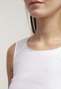 Vrouw in een mouwloos wit topje, met zichtbare nek, schouders en een deel van haar lippen tegen een effen lichte achtergrond.