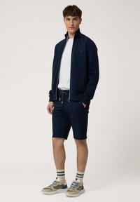 Chaqueta de fondo marino con cierre y cuello alzado, combinada con pantalones cortos de fondo marino. El modelo lleva una camiseta blanca y zapatillas beige con detalles azules y calcetines de rayas.