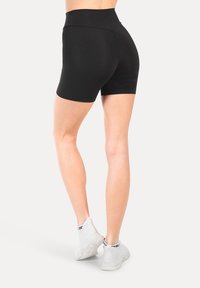 Pantaloni corti neri a vita alta realizzati in tessuto elastico, con una texture liscia e design aderente, abbinati a scarpe da ginnastica bianche.