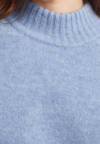 Pull bleu clair côtelé avec un col montant et un tissu doux et texturé. Présente un motif subtil et uniforme sur toute la surface.