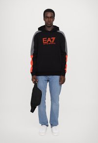 Felpa nera con maniche grigie e dettagli arancioni, dotata di una tasca frontale e il marchio EA7. Indossata con jeans chiari in denim e sneakers bianche.