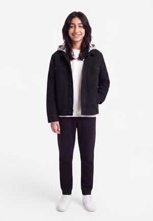 Adolescent aux longs cheveux foncés souriant, portant une veste noire avec capuche grise, chemise blanche, pantalon noir et baskets blanches, debout devant un fond blanc.