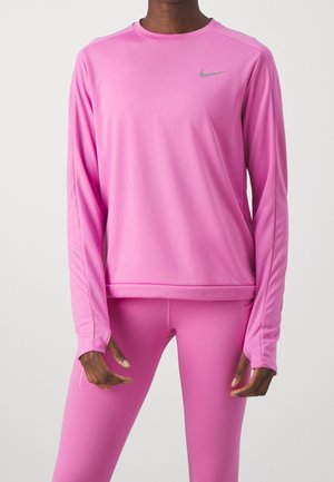 Osoba ubrana w różowy sportowy top z długim rękawem i dopasowane legginsy z małym szarym logo Nike na piersi na jednolitym tle.