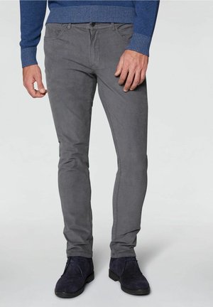 Man draagt strak zittende grijze corduroy broek, blauwe trui en donkerblauwe suède veterleren schoenen, staand tegen een effen achtergrond.