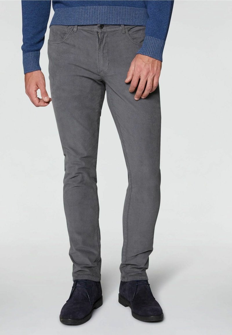 Homme portant un pantalon en velours côtelé gris slim, un pull bleu et des chaussures à lacets en suède bleu foncé, debout devant un fond uni.