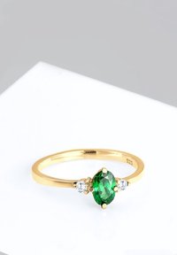 Bague en or avec une pierre précieuse ovale verte au centre, flanquée de deux petites pierres rondes transparentes sur une surface blanche.