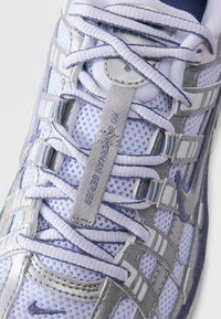 Primo piano di una sneaker Nike bianca e argento con tessuto a rete, lace intricati e etichetta "NIKE BY ERIC KAY SERIES" sulla linguetta.