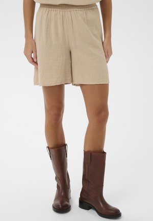 Beige teksturerede shorts med elastisk taljebånd båret med brune læderstøvler til midt på læggen på en stående model mod hvid baggrund.