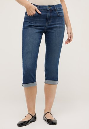 Femme portant un jean court bleu foncé retroussé et des chaussures Mary Jane noires, debout avec une main dans la poche.