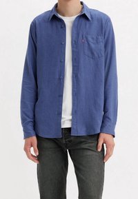 Niebieska koszula na guziki wykonana z miękkiego materiału, z kieszenią na lewej piersi i klasycznym kołnierzykiem. Noszona na białej koszuli.