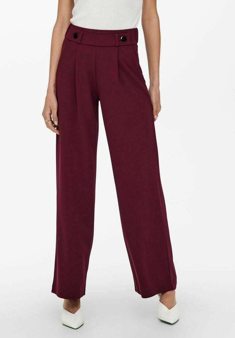 Femme portant un pantalon bordeaux taille haute à jambes larges avec des boutons noirs, associé à des chaussures blanches à bout pointu et un haut blanc sans manches.