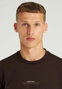 Bruine sweatshirt met een ronde hals en geribde manchetten, beschikt over witte logo tekst op de borst, gladde textuur, minimalistisch ontwerp.