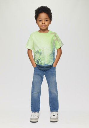 Grünes Grafik-T-Shirt mit einem Design, das Maschinen darstellt, kombiniert mit blauen Jeans und weißen Sneakers mit grauen Akzenten.