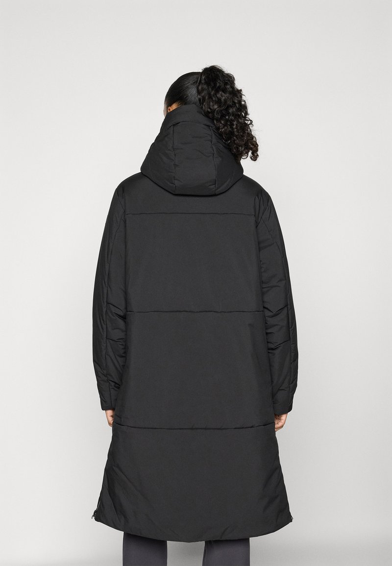 Frau mit lockigem Haar in einem Pferdeschwanz, die einen langen schwarzen gepolsterten Mantel mit Kapuze trägt, von hinten vor einem schlichten Hintergrund betrachtet.