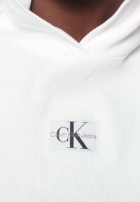 Biała bluza z kapturem o prążkowanej fakturze, z prostokątną metką z napisem "Calvin Klein Jeans" i stylizowanym logo "CK".