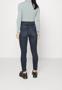 Donna con jeans skinny a vita alta blu scuro, maglione a maniche lunghe blu chiaro a costine e scarpe con piattaforma nere, in piedi su un pavimento grigio.