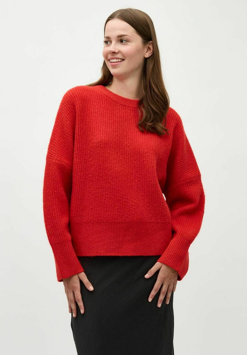 Roter gestrickter Pullover mit lockerer Passform, gerippter Textur und langen Ärmeln. Kombiniert mit einem schwarzen Rock, vor einem neutralen Hintergrund.