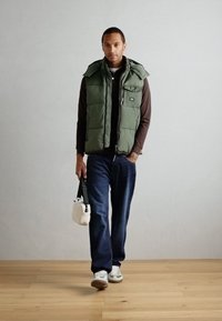 Green padded vest over a brown long-sleeve shirt, blue jeans, and white sneakers. Holds a small white bag with a black strap.