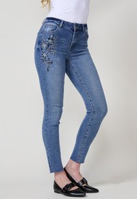 Jean slim en denim bleu clair avec broderies florales sur la cuisse gauche. Il dispose d'une taille standard et d'une coupe ajustée jusqu'à la cheville.