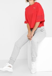 Czerwony krótki t-shirt z złotym logo, zestawiony z jasnoszarymi spodniami dresowymi z mankietami u kostek oraz białymi trampkami z wycięciem.