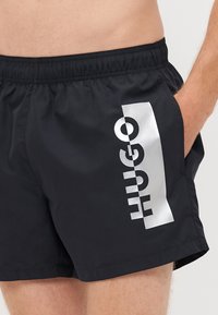 Shorts de bain noirs en tissu léger, arborant un logo "HUGO" blanc brillant sur le côté et une taille élastique pour plus de confort.