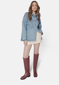 Denim jacket with a relaxed fit, light blue color, and button closure. Model wearing a cream pleated skirt and tall burgundy boots with a textured finish.