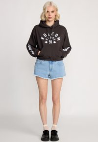 Un sweat à capuche gris foncé avec un logo blanc, une poche kangourou et des détails graphiques sur les manches ; associé à un short en denim léger effiloché et des chaussures noires.