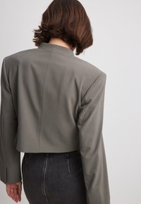 Blazer grigio accorciato con maniche lunghe, texture liscia e design minimalista. Presenta un colletto a scollatura e linee pulite sul retro.