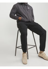 Grijze rits hoodie met een hoge kraag, zwarte cargobroek met elastische boorden en beige suède schoenen. De broek heeft zijzakken en een gestructureerde afwerking.
