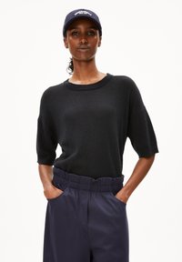 Schwarzer gerippter Pullover mit kurzen Ärmeln, kombiniert mit hochgeschnittenen navy Bl trousers, die einen elastischen Bund und seitliche Taschen haben.