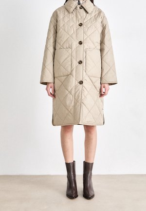 Manteau beige matelassé jusqu'aux genoux avec boutons sur le devant et grandes poches, porté avec des bottines marron foncé sur un fond neutre.